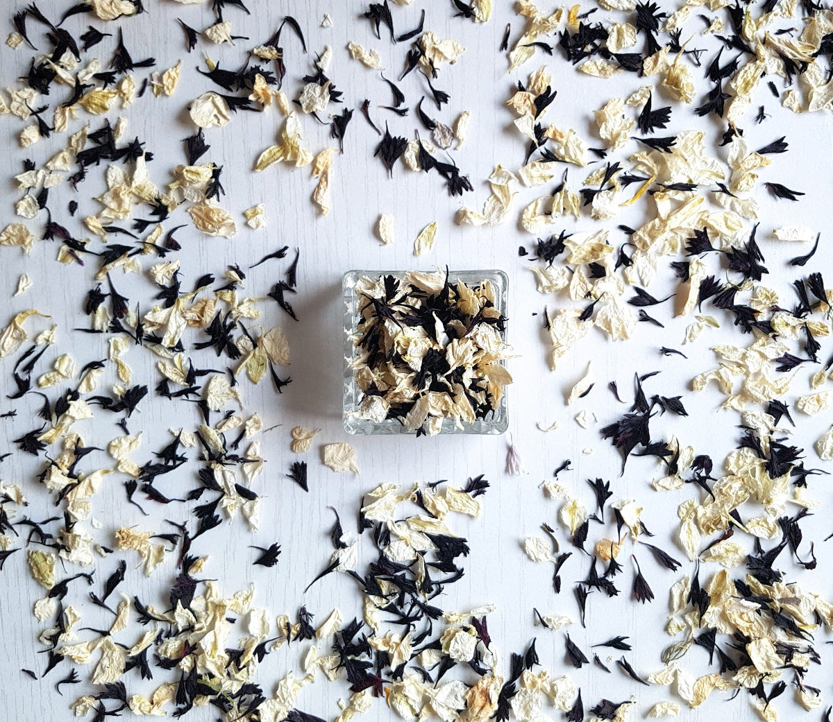 A small square glass container filled with dried flower petals, placed on a white surface and scattered petals