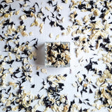 A small square glass container filled with dried flower petals, placed on a white surface and scattered petals